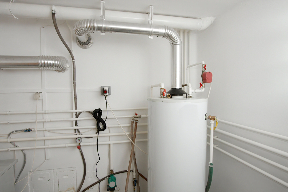 A water heater tank is installed in a utility room with multiple pipes, vents, and a control switch on the wall.