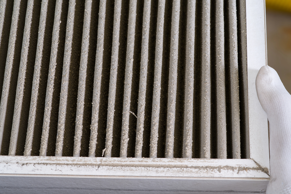 A close-up of a dirty air filter with dust buildup, held by a person wearing a white glove.