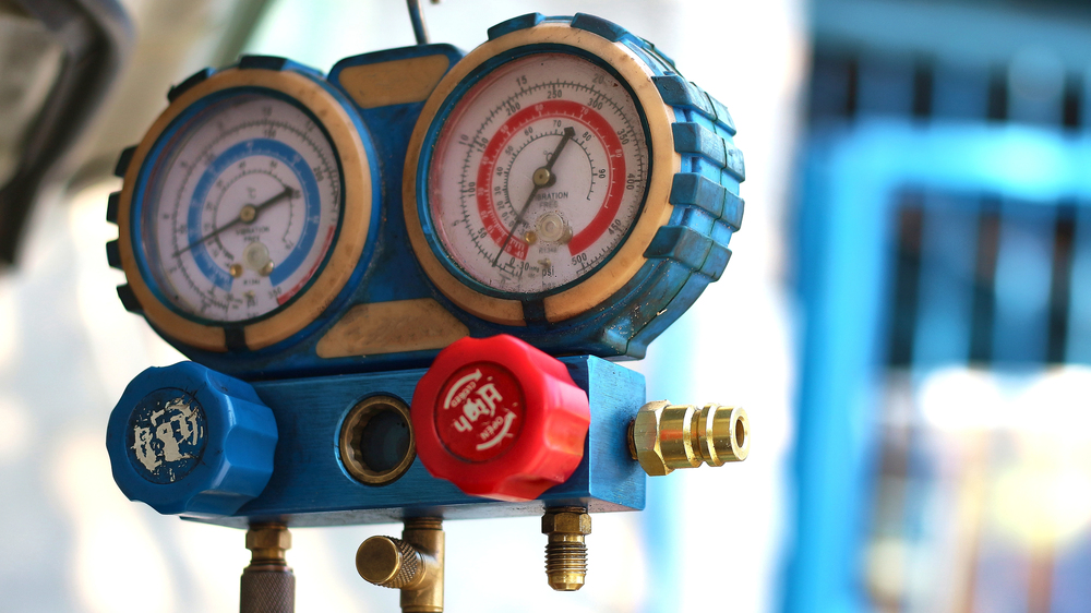 Close-up of a blue HVAC manifold gauge set with two pressure dials and red and blue valve knobs, used for checking refrigeration or air conditioning systems.