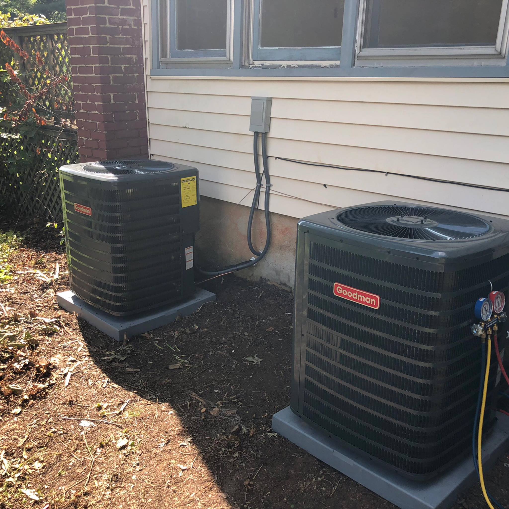 Two Goodman central air conditioning units are installed outside next to a house with siding. One unit has attached gauges for servicing.