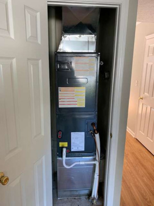 A residential HVAC furnace unit installed in a narrow closet space with the door open, showing ductwork and a white drain pipe below.