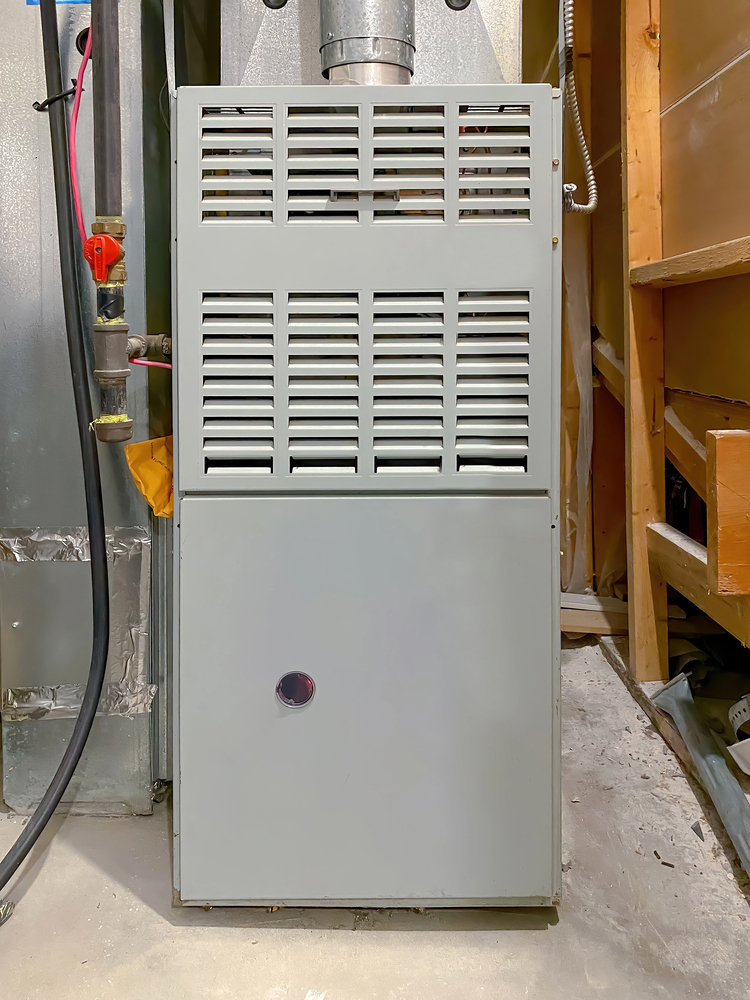 A gray metal furnace unit with a vented front panel is installed in a basement or utility room, surrounded by pipes and unfinished wooden walls.