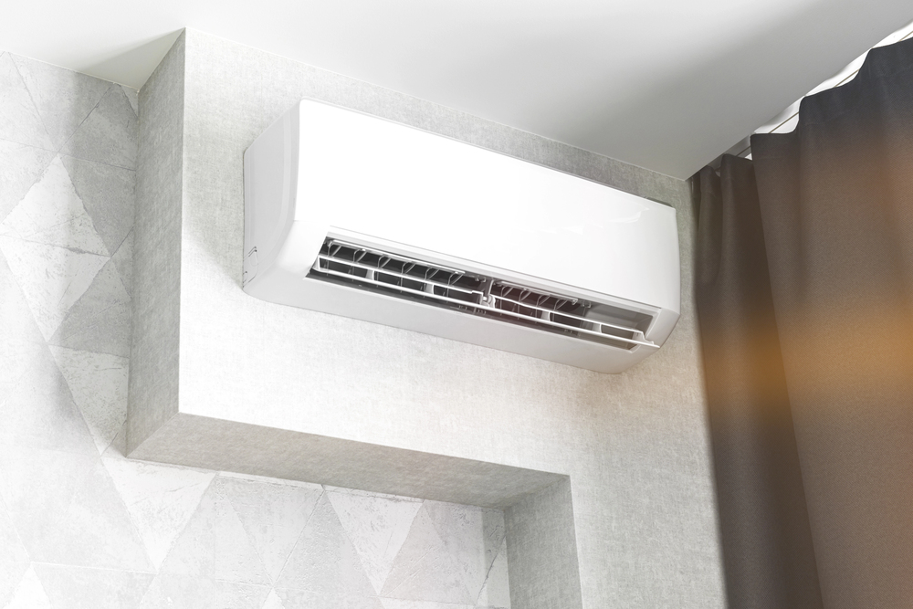 A white wall-mounted air conditioner is installed near the ceiling in a modern interior with geometric-patterned wallpaper and a dark curtain.